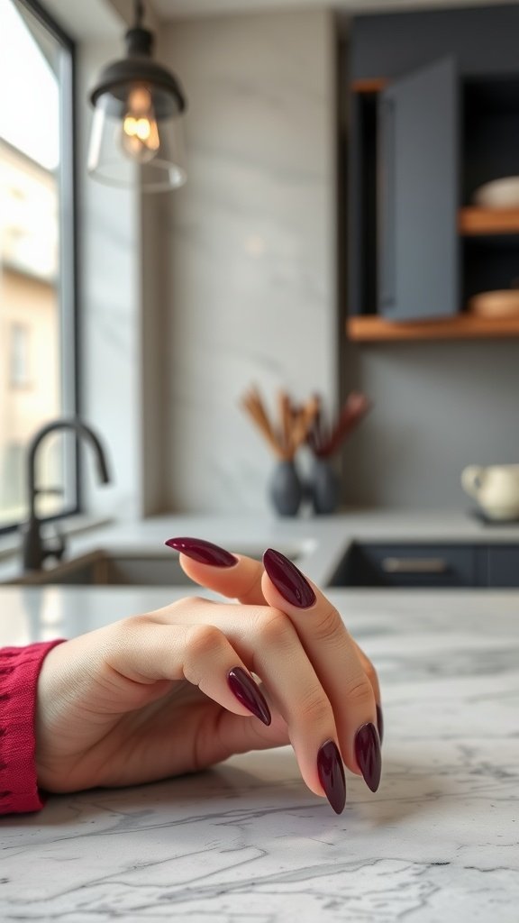 A close-up of a hand with half-moon cherry wine nails resting on a marble table.